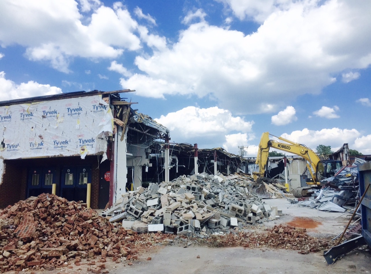 Yellow excavator demolishing a low-rise commercial building, with brick and concrete block debris in the foreground
