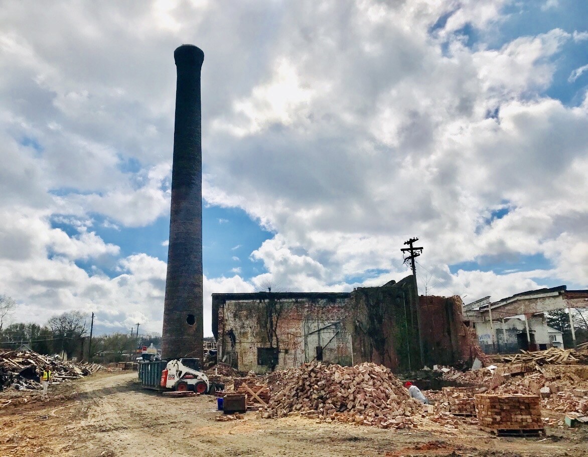 Industrial demolition site with brick rubble, heavy equipment, and a tall smokestack